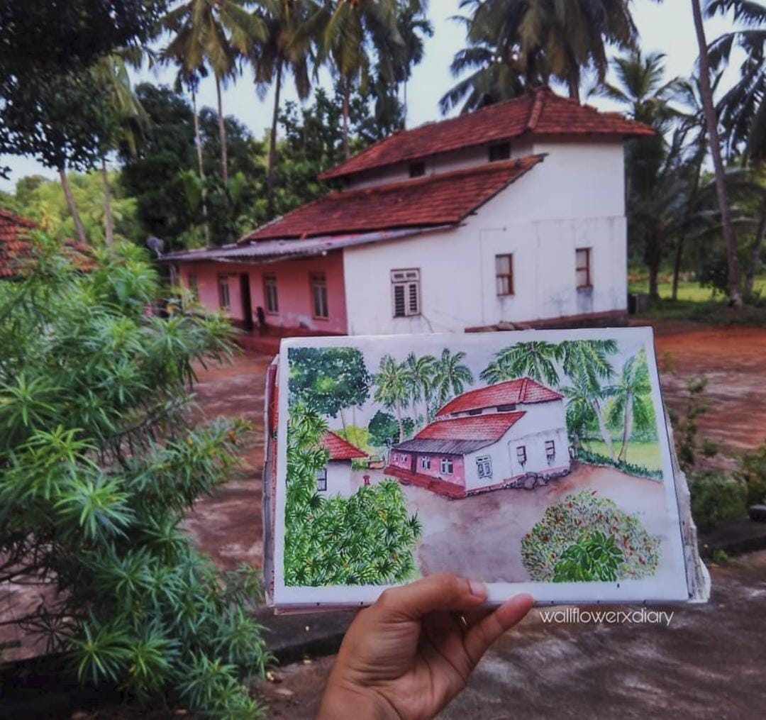 A watercolour painting of a tiled roof house held up so it lines up with the real house behind it.