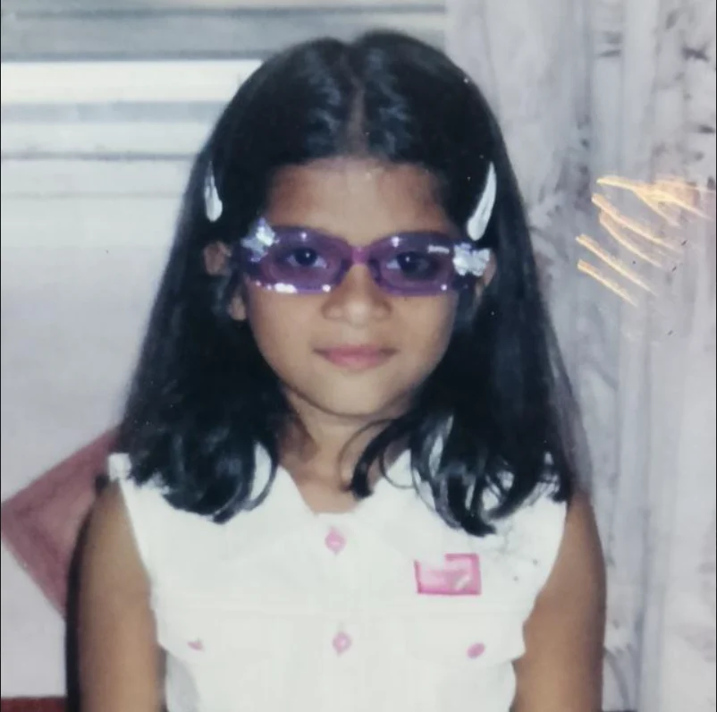 Theertha as a child, in purple sparkly glasses and a white top, looking straight at the camera.